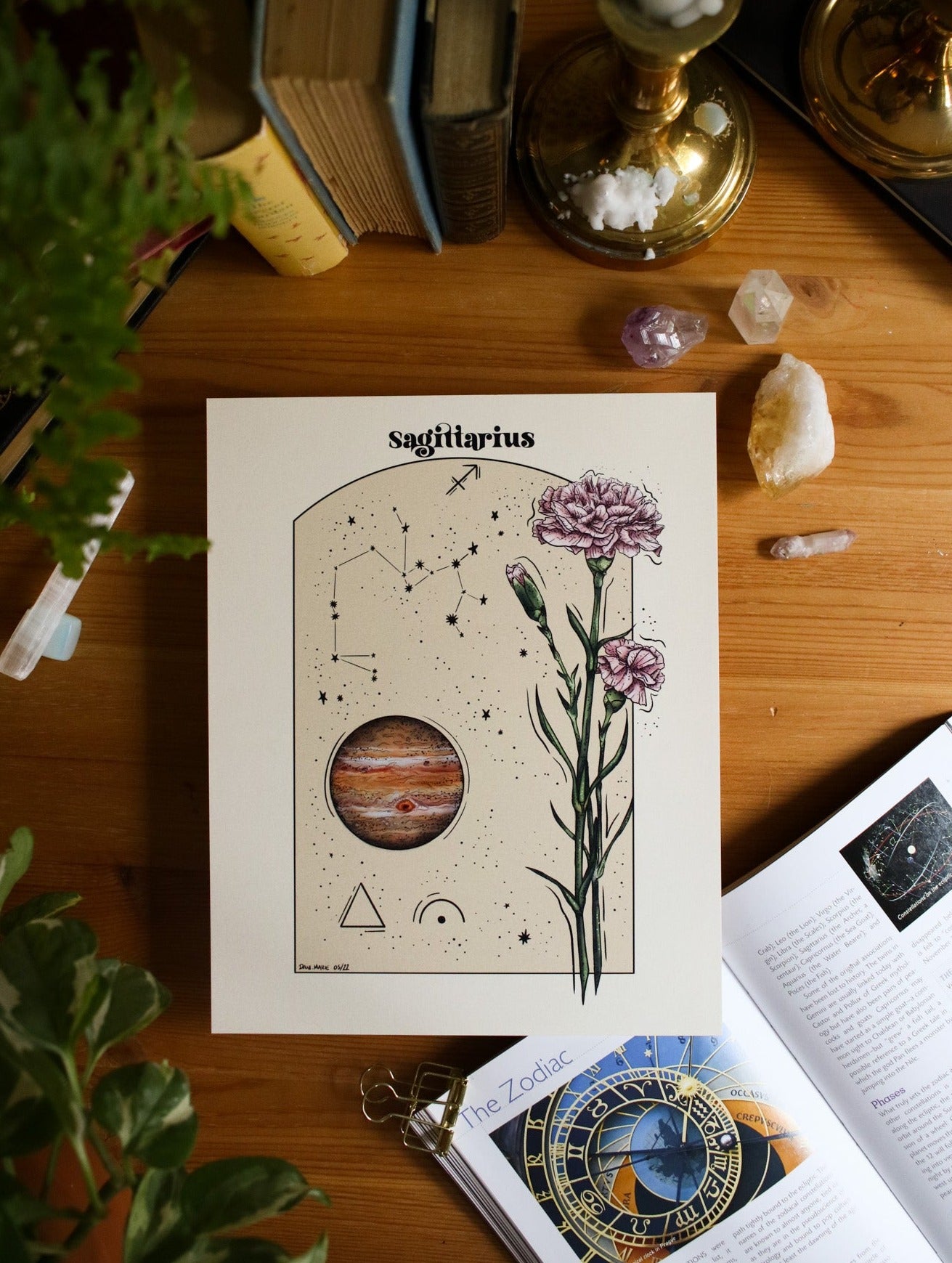Top down view of a piece of artwork of the sagittarius astrology sign info. The sag constellation, the planet Jupiter, pink carnations and a triangle for fire and an arch with a dot for mutable